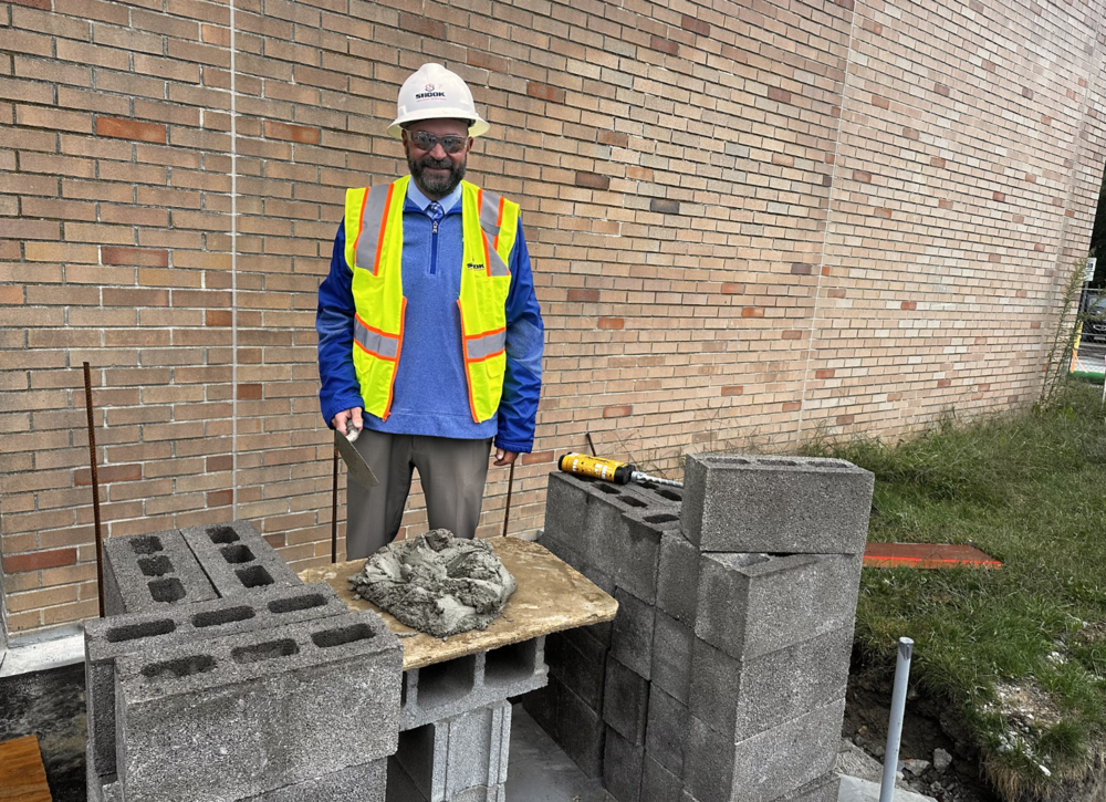 A man in a hard hat and yellow safety vest is standing with a trowel and cement on a construction site.