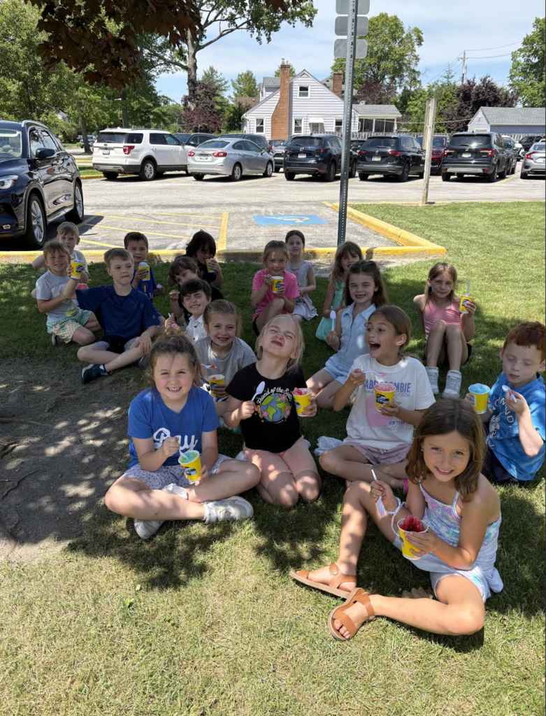 First-graders enjoy Kona Ice on the last day of school before summer break. Students sitting under a tree with an icee.