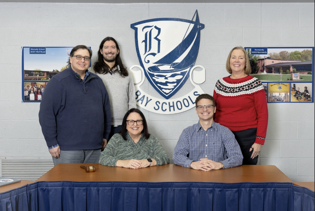 Board group photo 2026; five Board Members sitting and standing around a table with the district logo in the background.