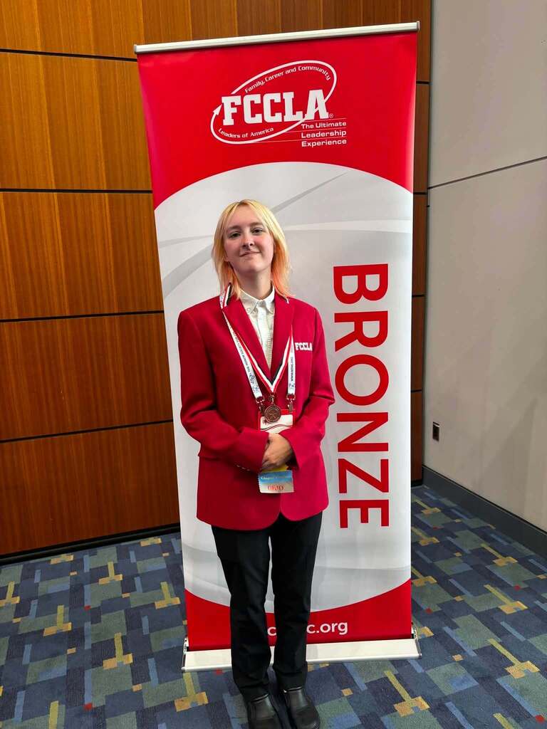 Photo shows a blonde young women standing in front of a vertical banner and wearing a medal around her neck. She has on a red jacket.