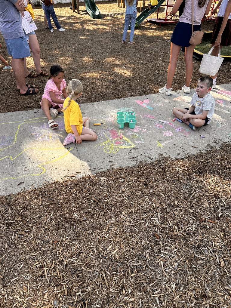 Three kindergarten students sit on a sidewalk chalking the walk at their new school.