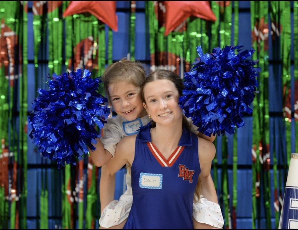 A photo of a young girl with blue pom poms on the back of a high school cheerleader in a blue uniform.