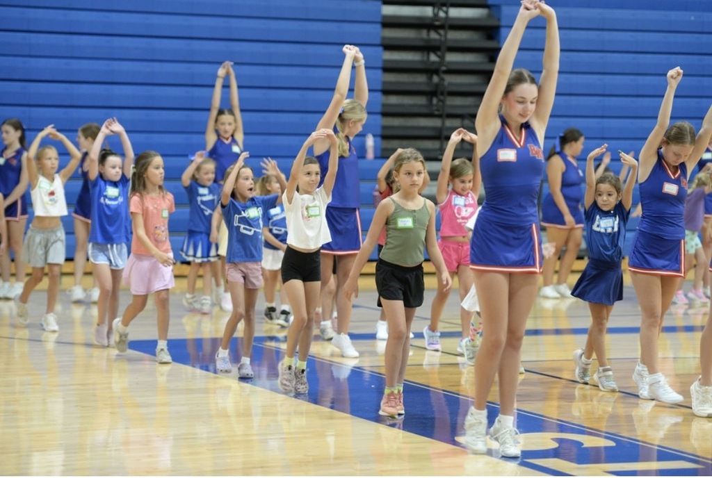 A photo of high school cheerleaders leading young students in learning a cheer in a gym.