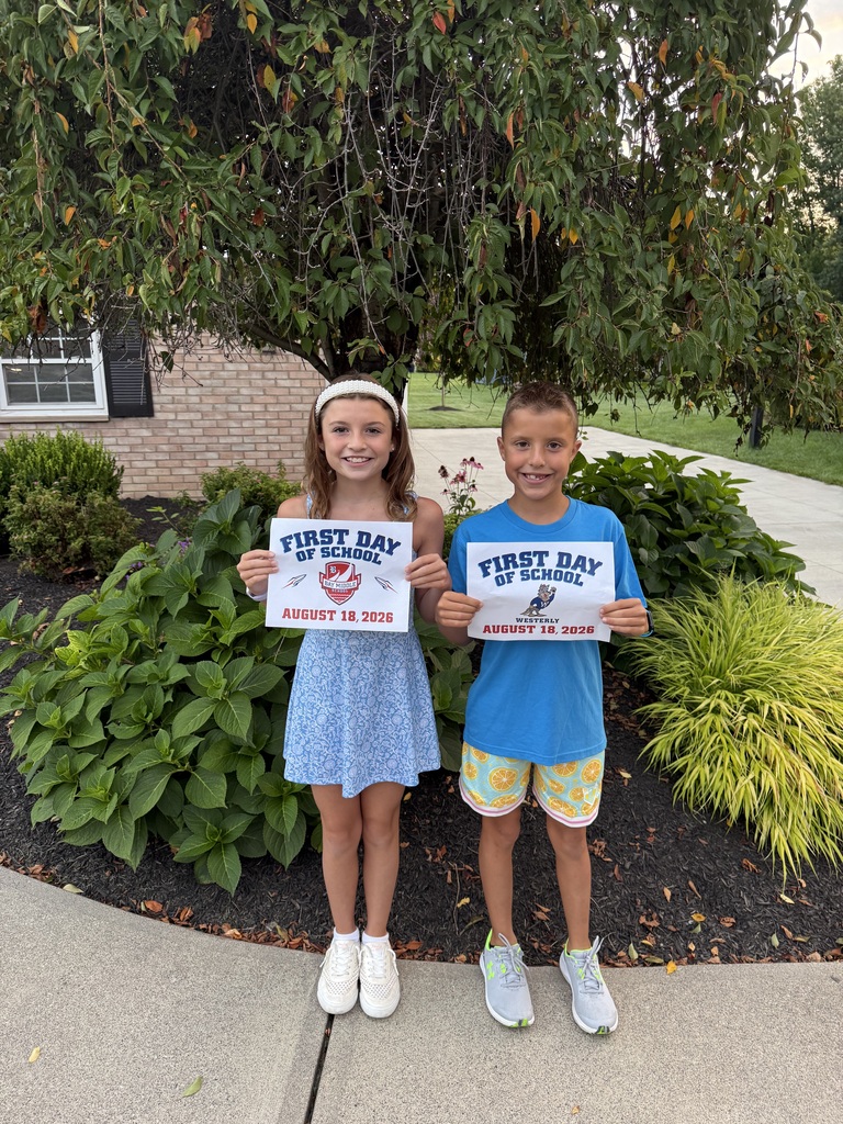 Two students standing in front of shrubbery on their first day of school, holding up signs that say that.