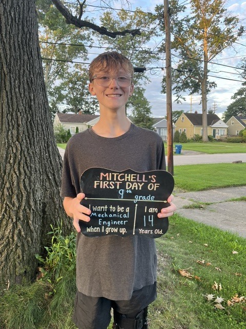 Photo of a freshman boy student on his first day of high school, holding a sign standing by a tree.