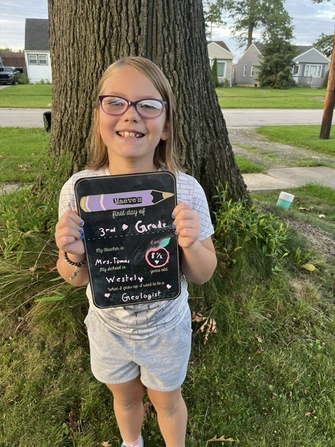 A female third-grader holding a first day of school sign standing in front of a tree. She has glasses and a blue outfit on.
