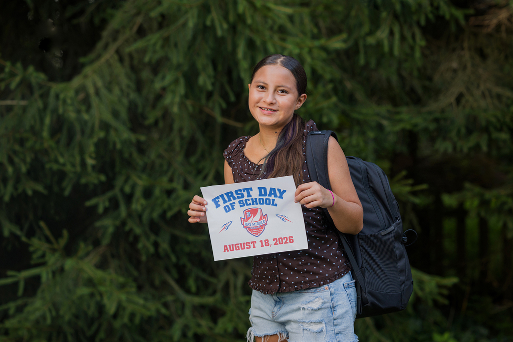 A middle school girl standing in front of a fir tree holding a first day of school sign.