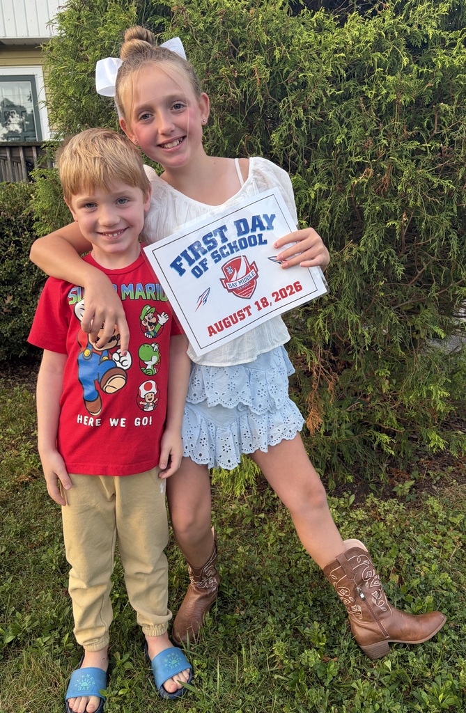 A middle school girl and her little brother standing in front of shrubbery and she's holding a back to school sign.