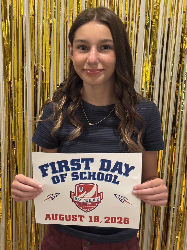 A middle school girl is standing in front of gold streamers holding a first day of school sign.