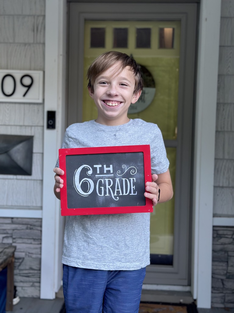 A middle school aged boy standing on his front porch holding a first day of school sign.