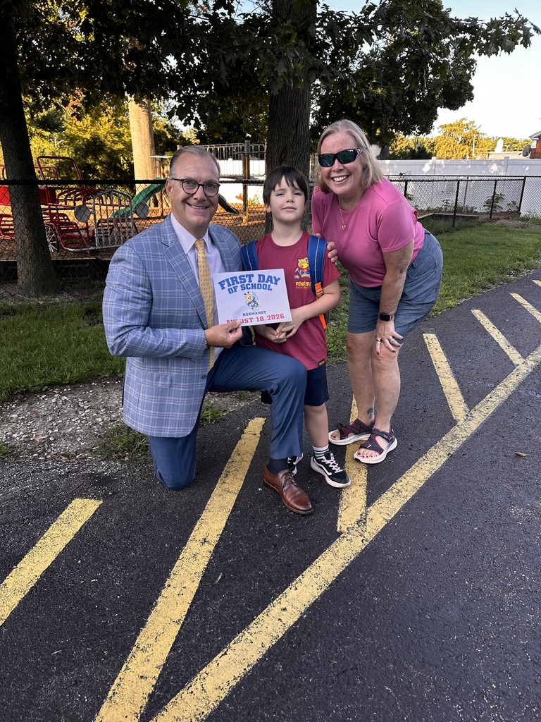 An elementary-aged boy flanked by the supt and his grandma, holding a first day of school sign.