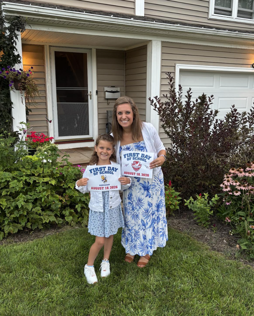 An elementary girl on the front lawn with her mom, both holding first day of school signs. 