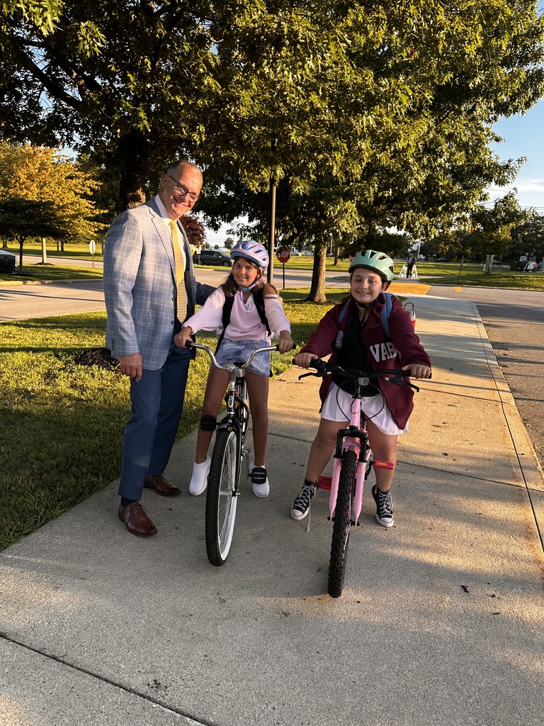 Two middle school girls on bikes pose for a photo with the superintendent on school campus.