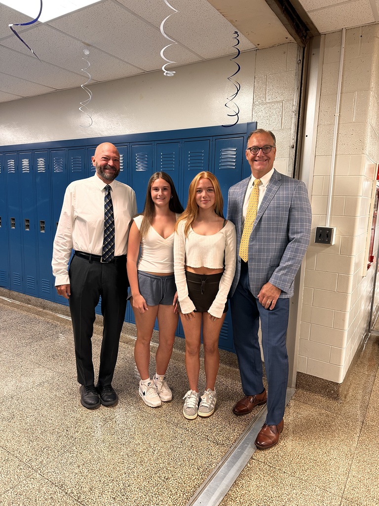 Two female high school students are flanked by their principal and their superintendent on the first day of school. They are standing in front of blue lockers. 