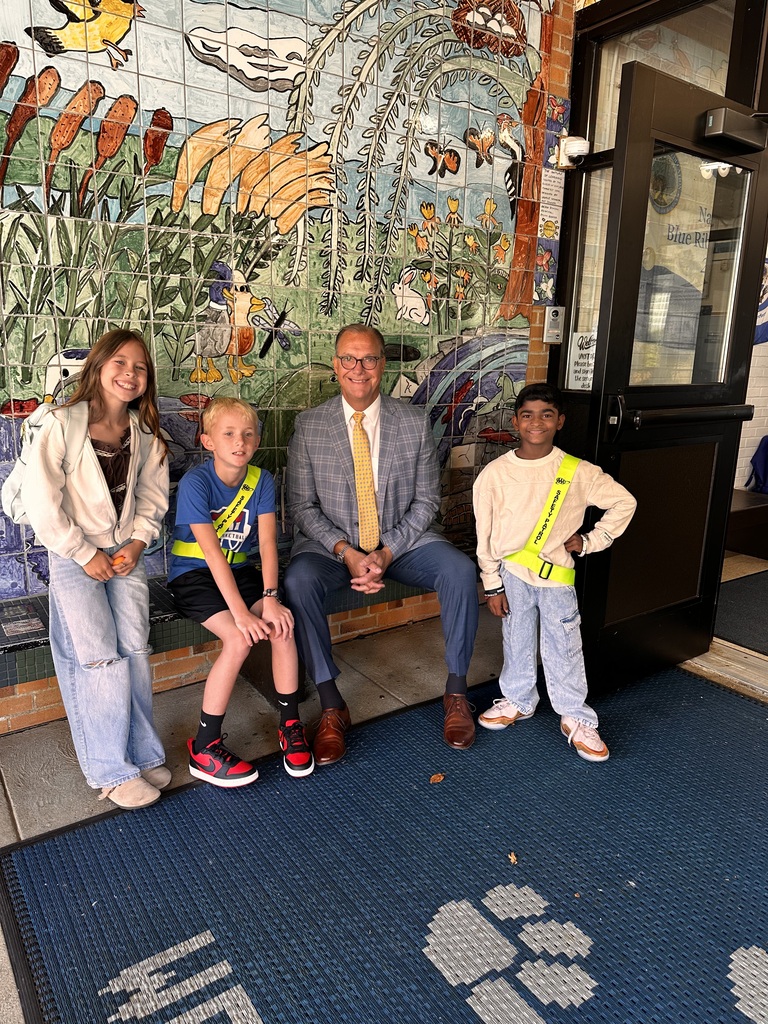 Three elementary students are with their school district superintendent, sitting and standing on a bench near the school entrance. Two of the students have safety patrol uniforms on.