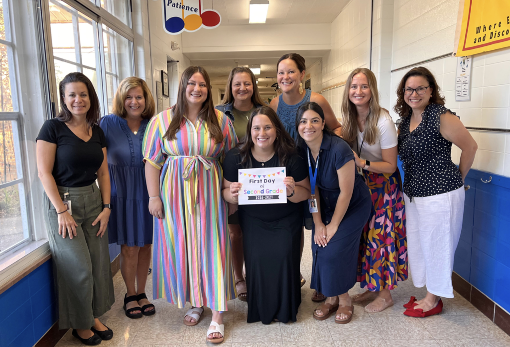 Nine second-grade teachers lined up in the hallway of their school with a first day of school sign.