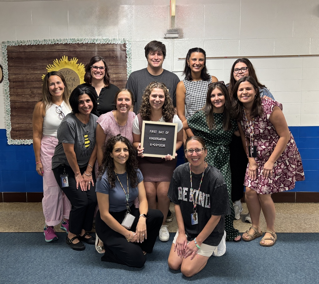 12 kindergarten teachers pose in the hallway of their school with a first day of school sign.