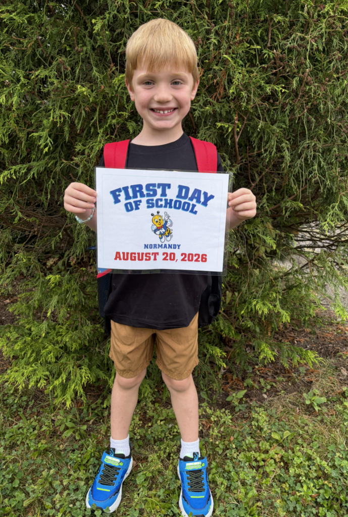First day of school photo with a kindergarten student standing in front of a pine tree holding a first day of school sign.