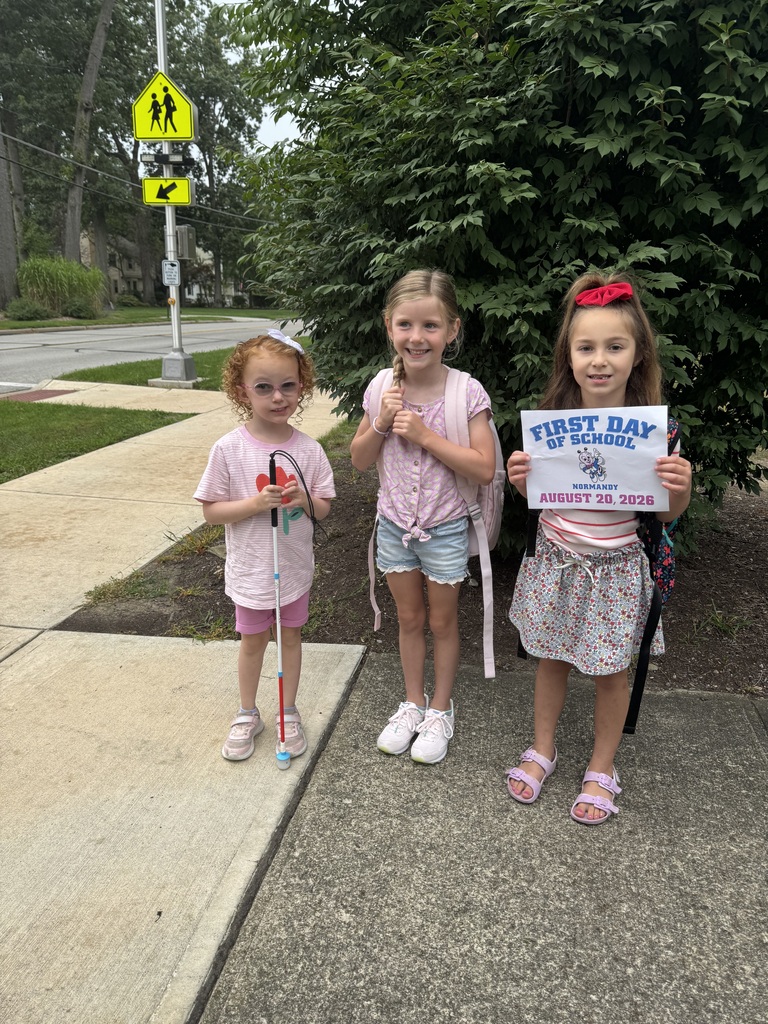 Three kindergarten girls ready for their first day of school standing in front of a shrub with a first day of school sign.