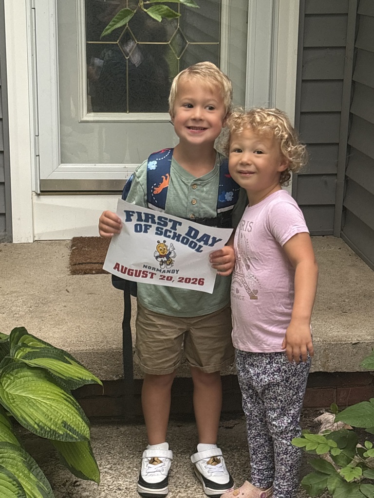 A kindergarten student on his front porch with his little sister holding a first day of kindergarten sign.