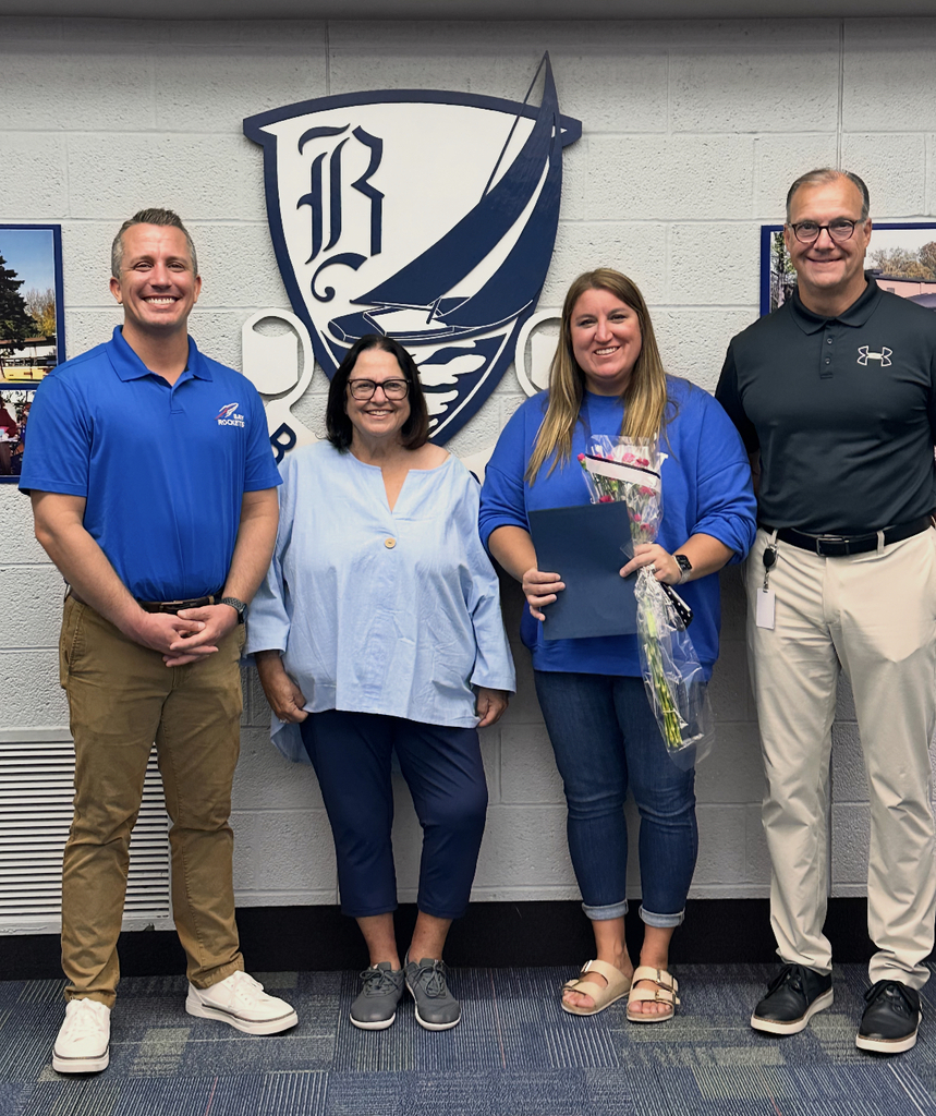 School administrators stand in front of the district's crest as the board president hands a certificate and flowers to the one winning the award.