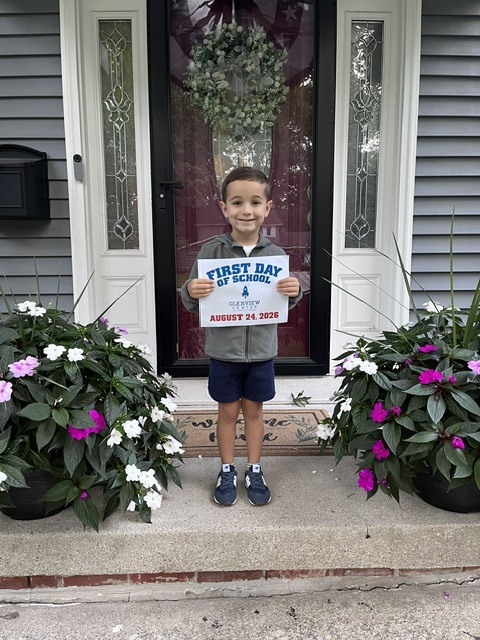 First day of preschool for this young man standing on his front porch holding a first day of school sign, flanked by two flower pots.