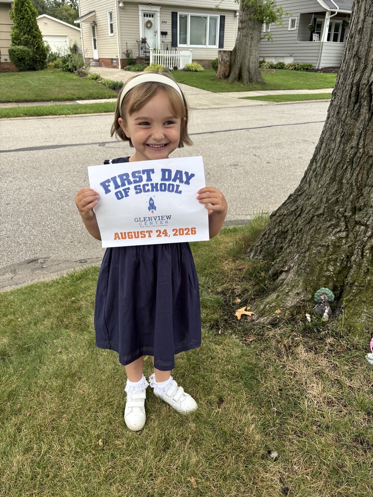 A little girl standing by a tree, dressed in blue and white and holding a first day of preschool sign.