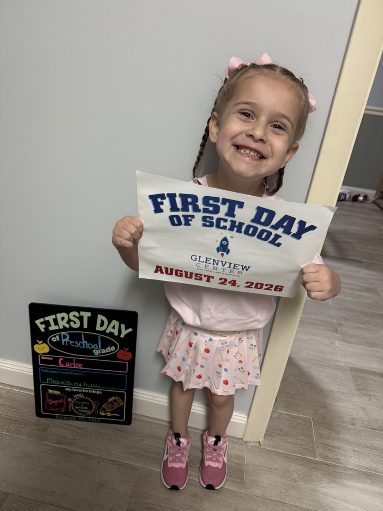 A little girl standing in front of a gray wall holding her first day of preschool photo. She is dressed in pink from hair bows to tennis shoes.