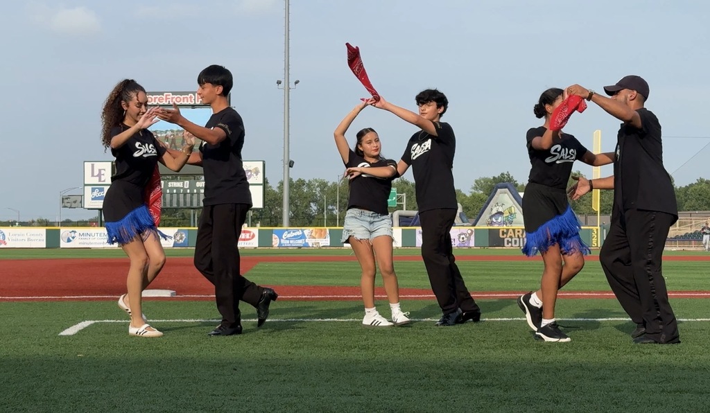 A middle school student and her dance partner salsa dancing at a practice outside on a baseball field.