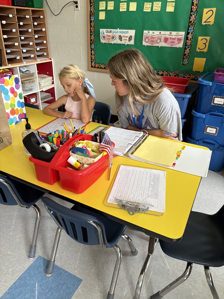 A high school female student works one-on-one with a first-grade female student as part of the educational aide experienceship program. They are seated in a classroom at a yellow table with small blue chairs.