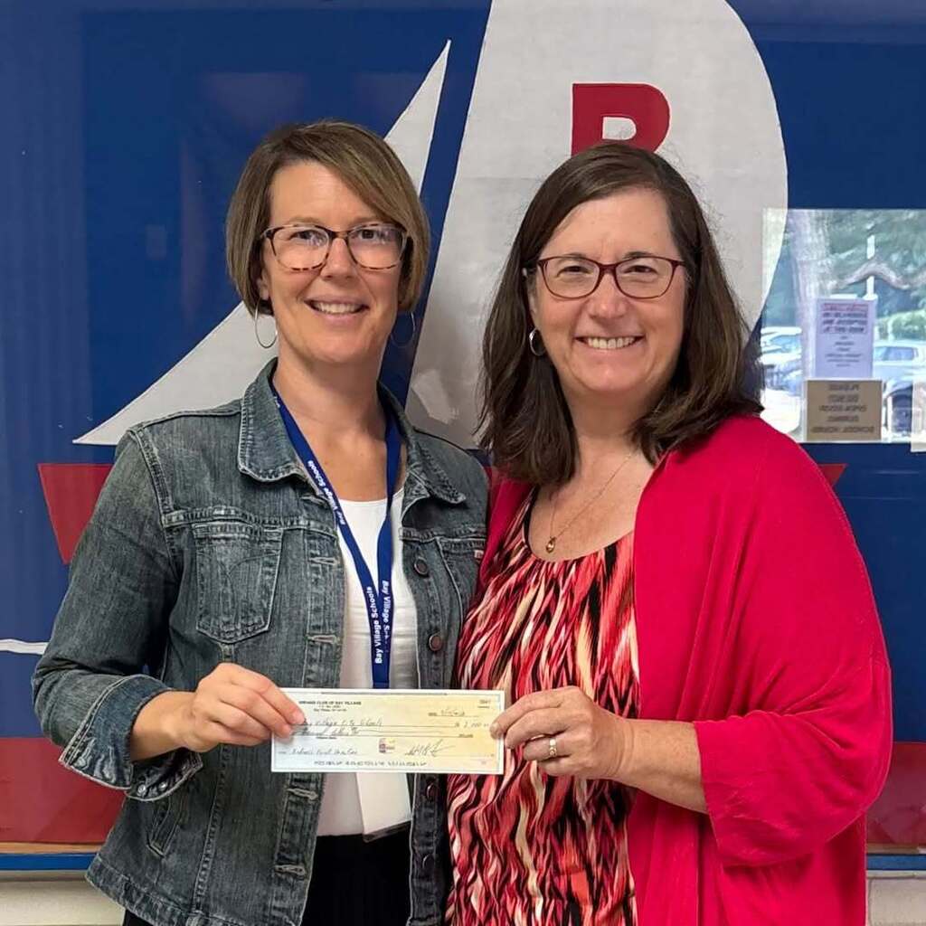 Two ladies smiling and holding a check in front of a red, white and blue background. The check is from Bay Kiwanis for Bay Village Schools.