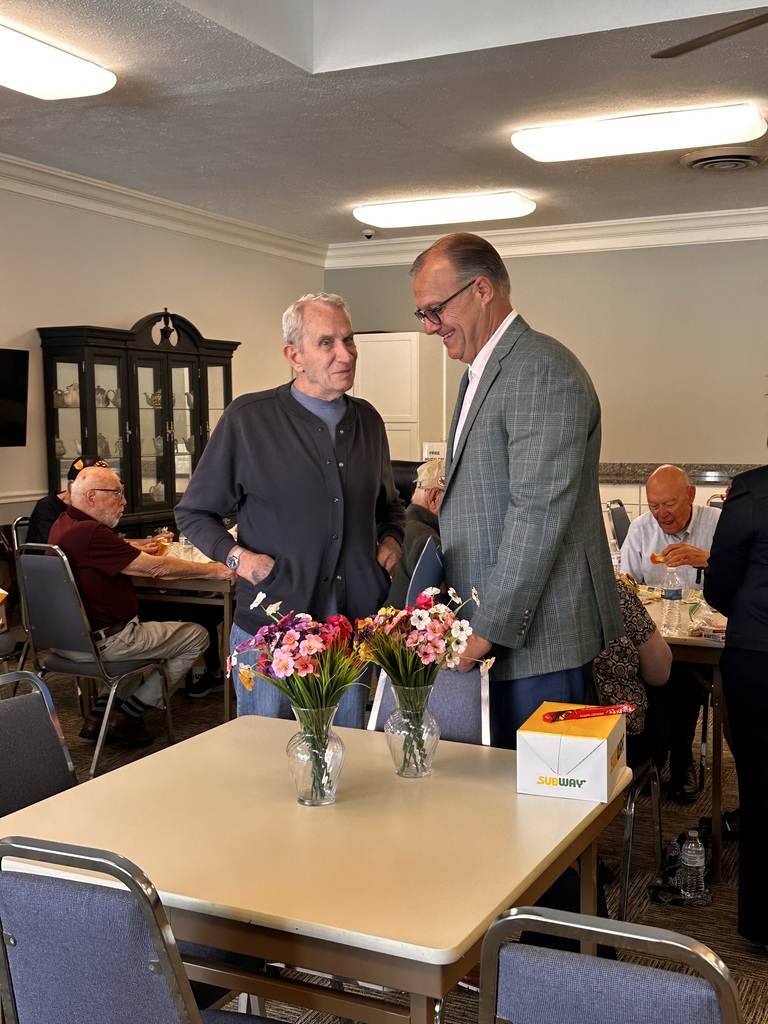 Two men, standing, share a conversation in the senior citizen hall while others are seated behind them eating lunch and playing games.