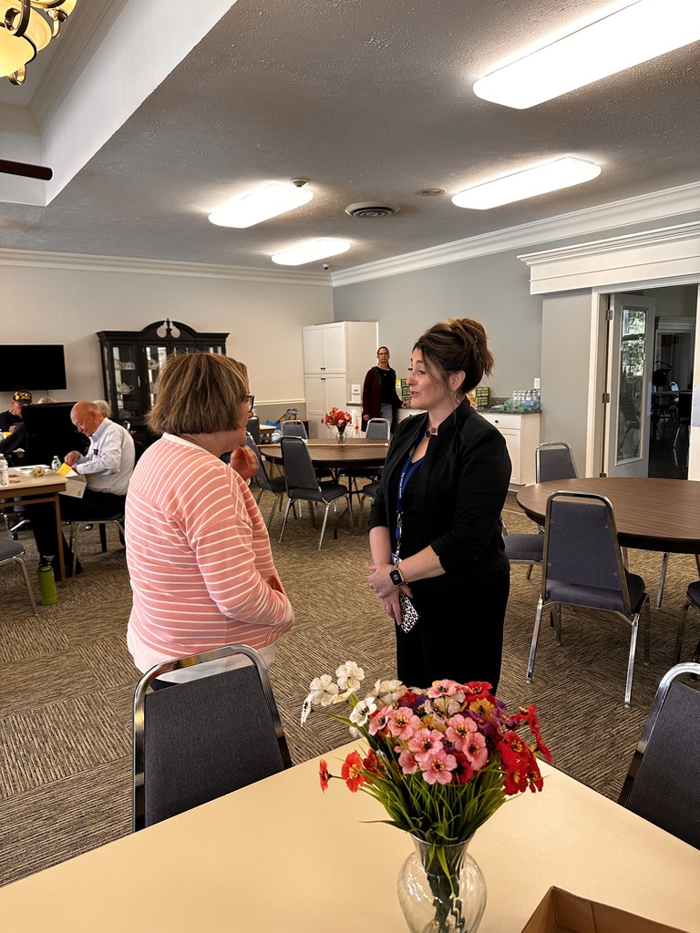 A woman talks to a senior citizen female, both standing, in a room full of round tables and chairs.