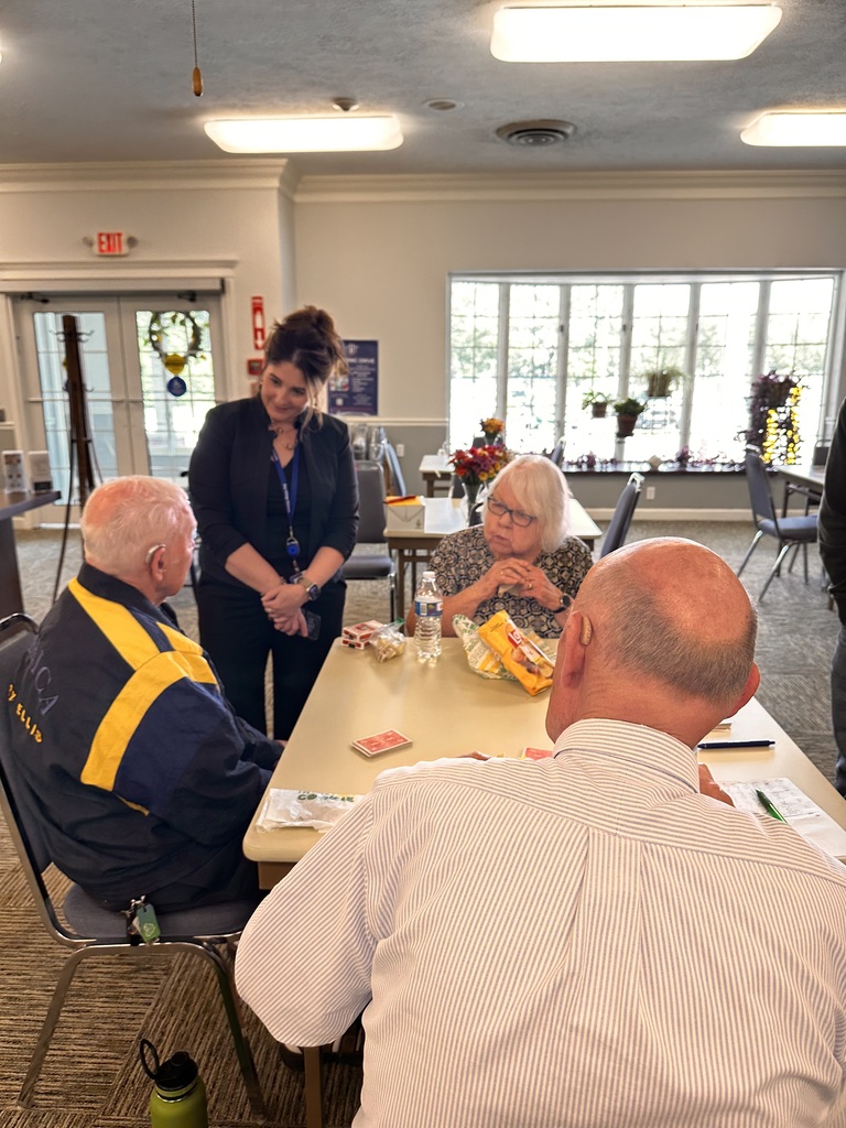 A woman is standing and chatting with a small group of senior citizens seated around a square table.