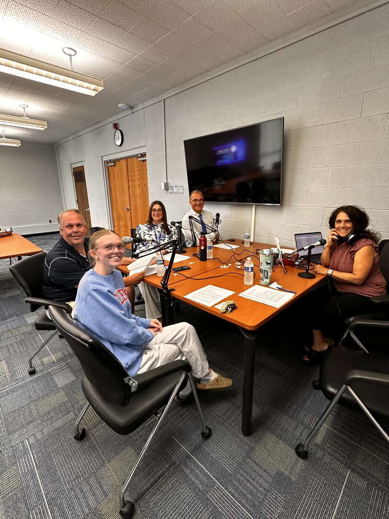 A photo of four adults and one student seated around tables as they record a podcast episode. There are mics and cords and notes on the table.
