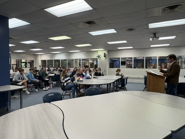 A man stands at the podium of a room full of seated high school students.