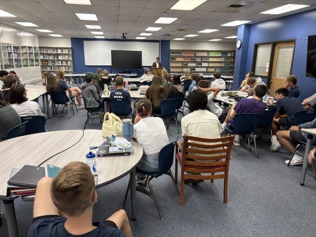 A man stands at a podium speaking to a group of seated high school students, talking about what it's like to be a sports reporter. 