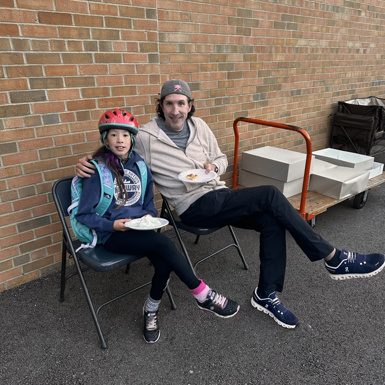 my dad and his daughter with bicycle helmets on sitting on a bench at an event at her school in the morningm