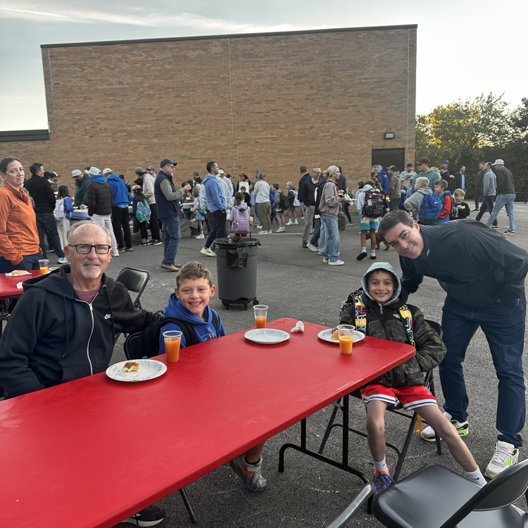 two dads in their son seated at a table having a doughnut at their children’s school.