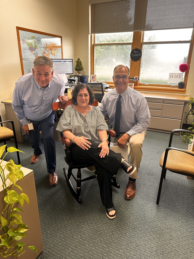 A principal and a superintendent flank a gift coordinator who is seated in a rocking chair in her office in the school building.