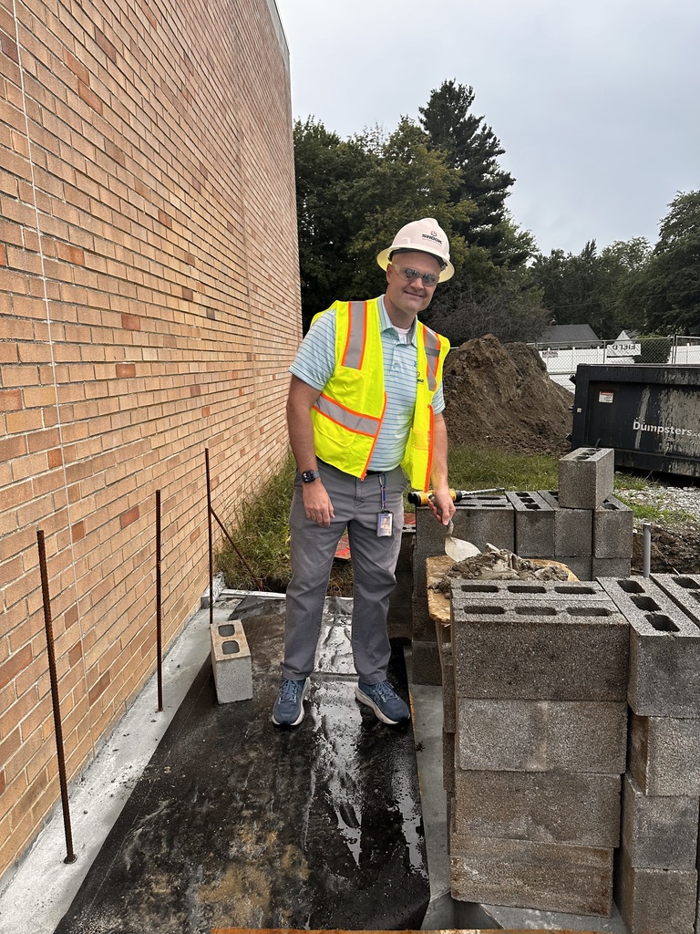 A man in a hard hat and yellow safety vest prepares a brick at a construction site.