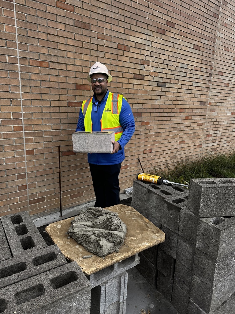 A man in a hard hat and yellow safety vest carries a brick at a construction site.
