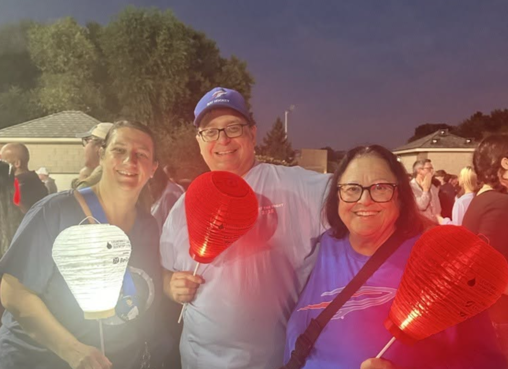 three adults holding lighted luminaria at dusk all wearing red, white and blue spirit wear