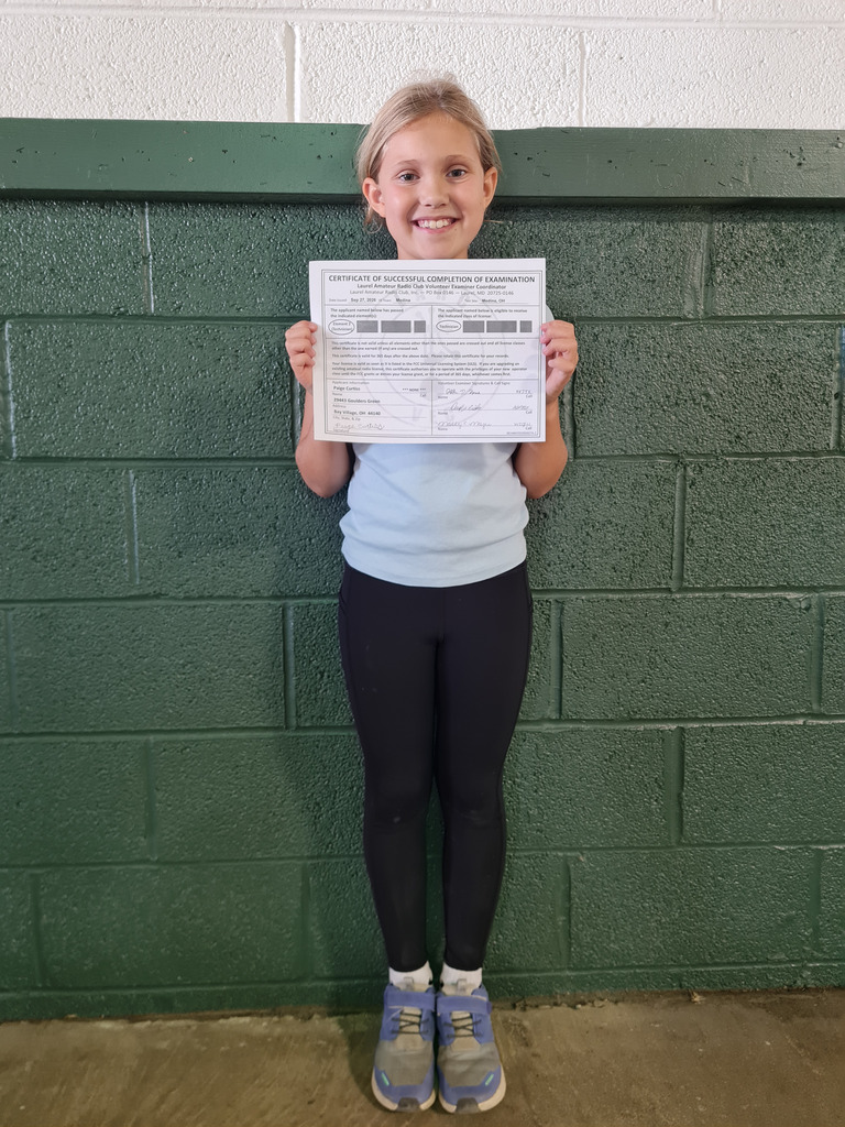 An elementary student holds her Ham Radio certificate, standing against a green brick wall.