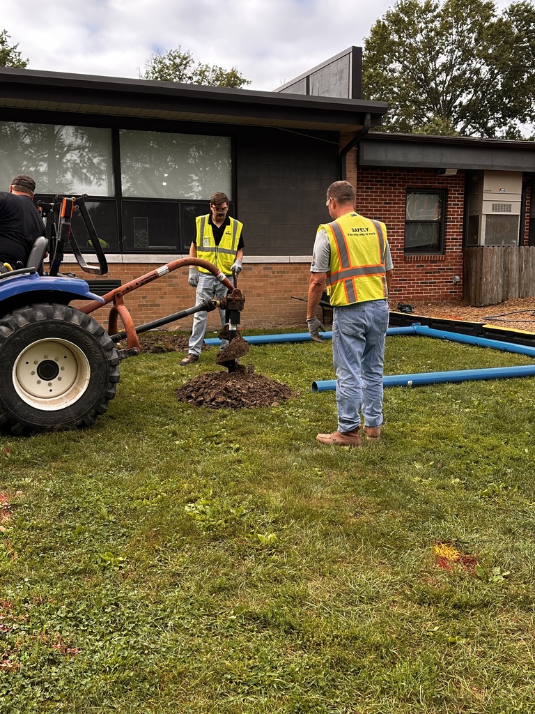Two men in yellow safety vests direct a backhoe tractor driver as they drill holes in the ground to install a blue swing set pole.