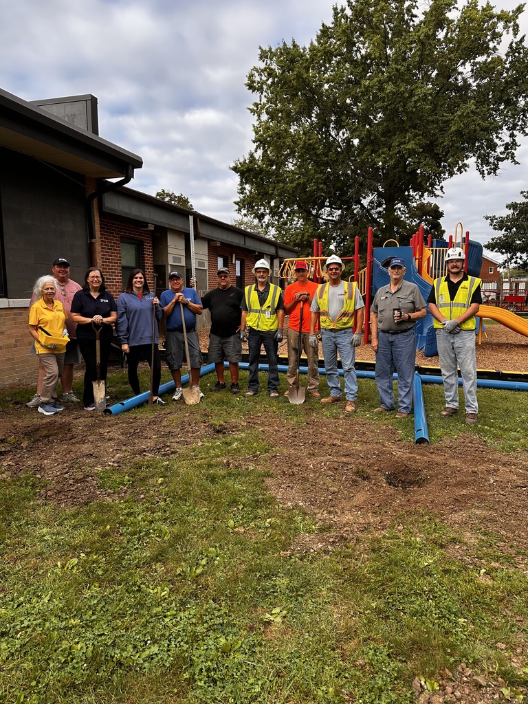 Eleven adults in hard hats, safety vests and with shovels, get ready to install a blue pole in the ground for a new adaptive swing set.