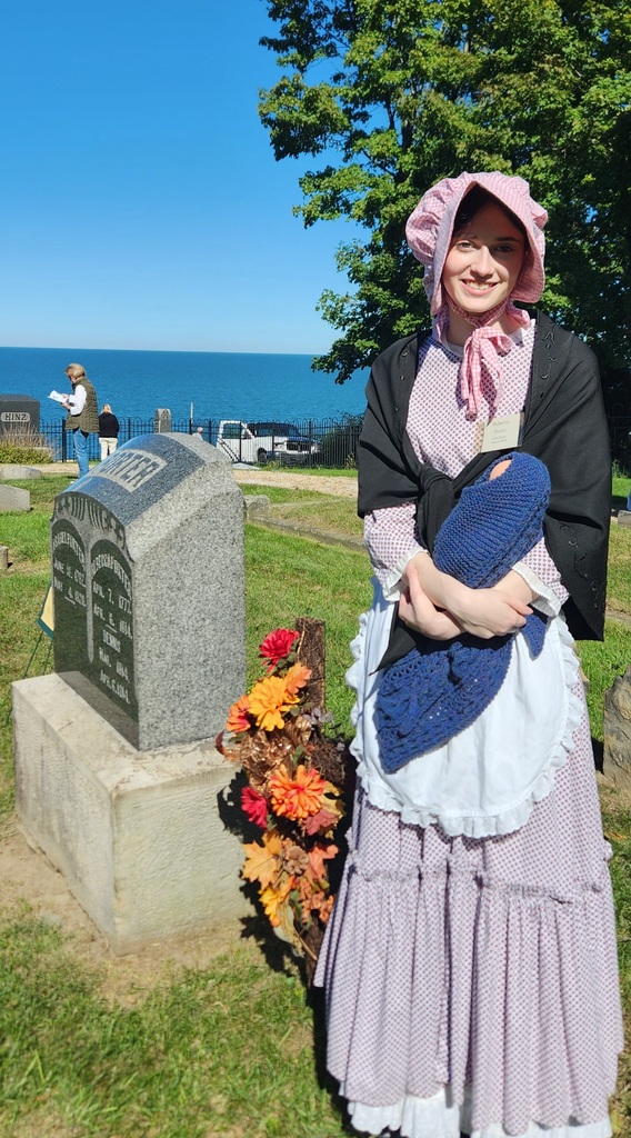 A student dressed in colonial period dress standing by a tombstone in a cemetery by the lake.