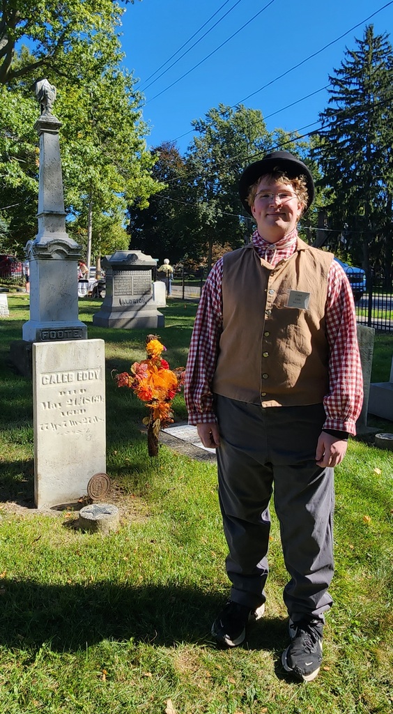 A student standing in colonial costume by a tombstone in a cemetery.