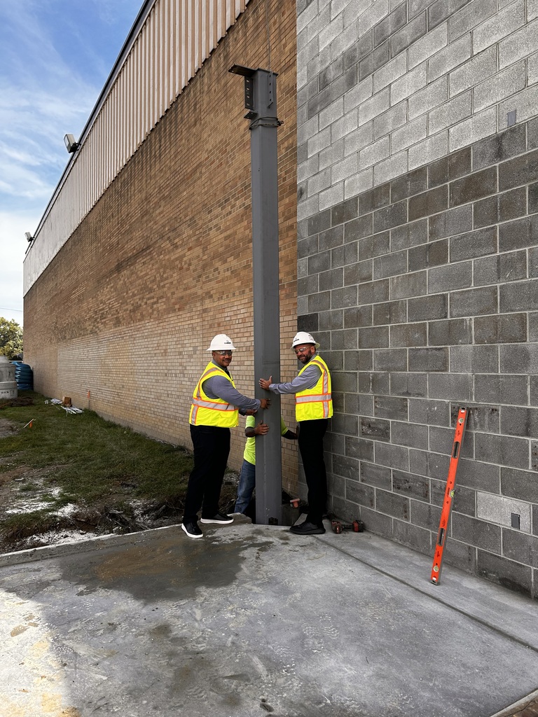 Two men in yellow safety vests and white hard hats install a beam footer into the ground outside a high school building.