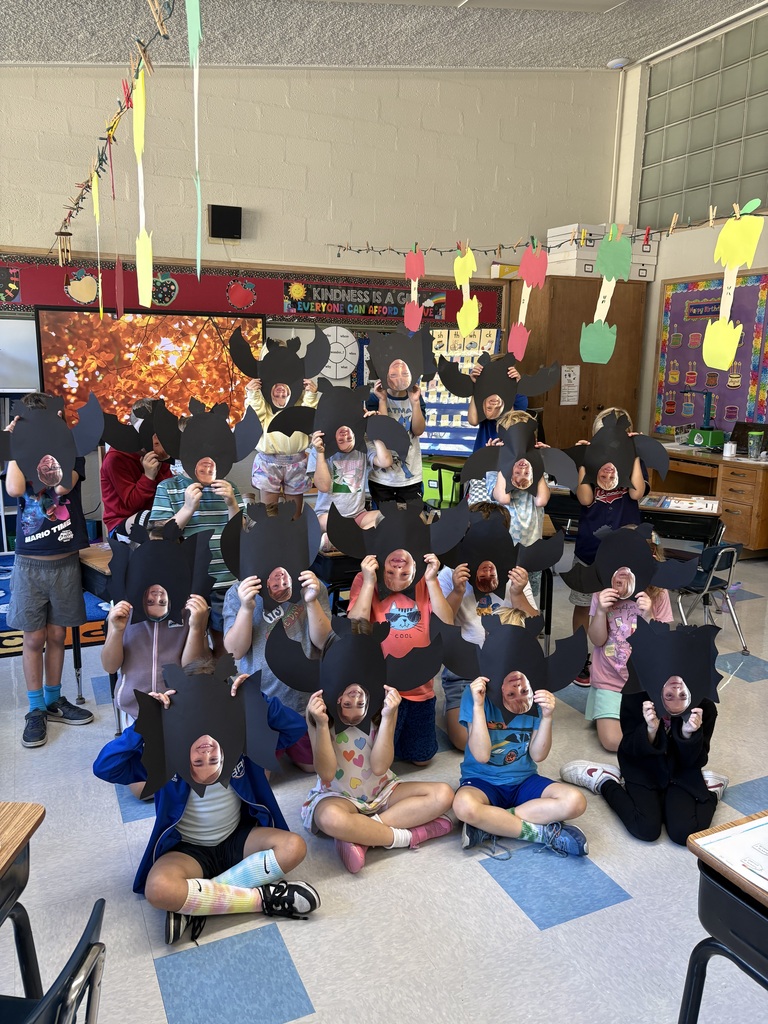 A group of first-graders posed for a photo in rows, some seated, some standing, each holding up a homemade black bat.